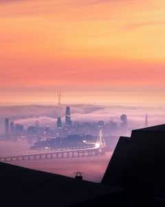 Vista panorámica de una ciudad envuelta en niebla durante el amanecer, con un cielo de tonos naranjas y morados.